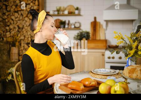 Frau trinkt Milch, während sie am Tisch in einer rustikalen Küche sitzt. Der Tisch wird mit frischen Brötchen, Äpfeln serviert und mit Wildblumen dekoriert Stockfoto