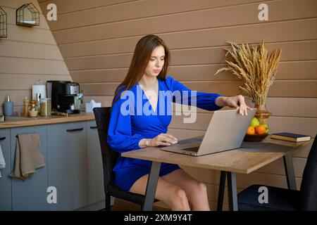 Eine fokussierte Frau sitzt an einem Holztisch in einer gut beleuchteten, modernen Küche und öffnet einen Laptop in der modernen Küche Stockfoto