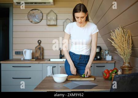 Frau steht in einer gut beleuchteten, modernen Küche und hackt Gurke auf einem hölzernen Schneidebrett Stockfoto