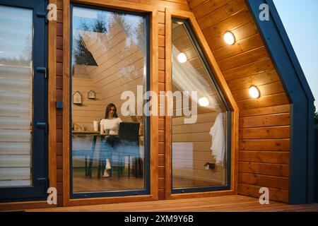 Blick durch große Glasfenster auf eine gemütliche, moderne Holzhütte auf eine Frau, die während der Dämmerung an einem Tisch sitzt Stockfoto