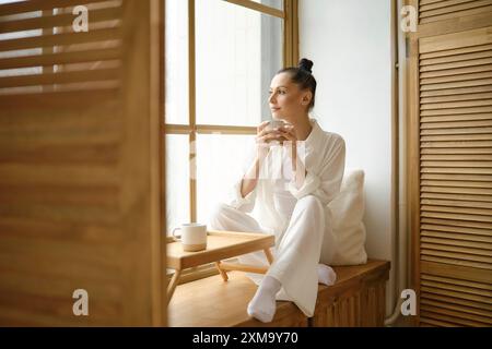 Traumhafte Frau in weißen Kleidern, die auf der Fensterbank sitzt und mit beiden Händen eine Tasse Kaffee oder Tee hält Stockfoto