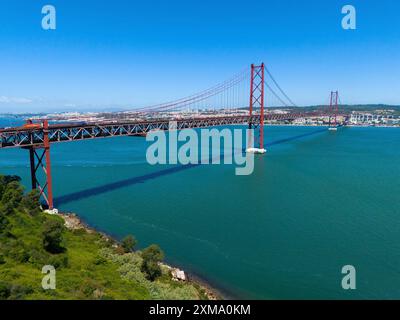 Eine große rote Brücke erstreckt sich über eine weite Wasserfläche unter einem klaren blauen Himmel, umgeben von grüner Landschaft, aus der Vogelperspektive, Ponte 25 de Abril Stockfoto