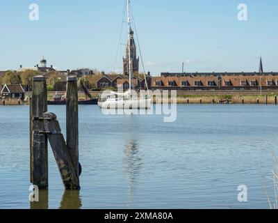 Ein weißes Segelboot auf einem Fluss vor einem Dorf, alte Holzpfosten im Wasser im Vordergrund, enkhuizen, niederlande Stockfoto