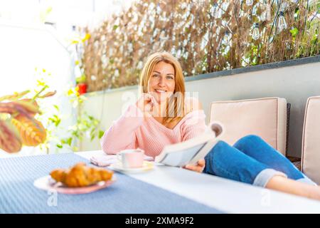 Erwachsene weiße blonde Frau lächelt in die Kamera und liegt auf einem Sofa im Garten, genießt Frühstück und liest ein Buch Stockfoto