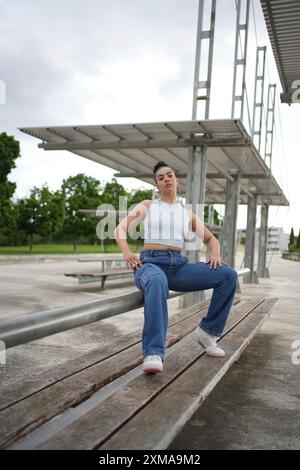 Eine Frau sitzt auf einer Bank im Park. Sie trägt blaue Jeans und ein weißes Tanktop. Die Szene ist friedlich und ruhig, mit Bäumen im Hintergrund Stockfoto