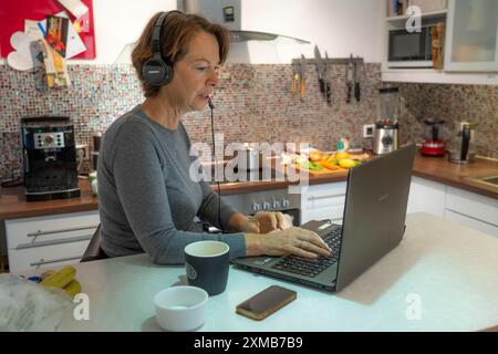Frau, Mitte 50, arbeitet von zu Hause aus, mit Laptop und kommuniziert mit Kollegen über Headset, Home Office oder in der Küche Stockfoto