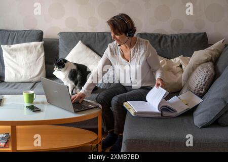 Frau, Mitte 50, arbeitet von zu Hause aus, mit Laptop und kommuniziert mit Kollegen über Headset, Home Office oder auf dem Sofa Stockfoto