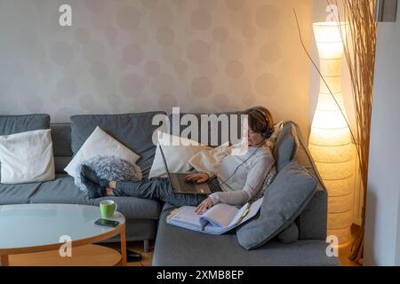Frau, Mitte 50, arbeitet von zu Hause aus, mit Laptop und kommuniziert mit Kollegen über Headset, Home Office oder auf dem Sofa Stockfoto