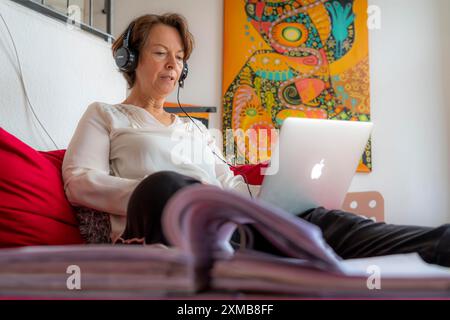 Frau, Mitte 50, arbeitet von zu Hause aus, mit Laptop und kommuniziert mit Kollegen über Headset, Home Office oder auf dem Sofa Stockfoto