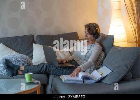 Frau, Mitte 50, arbeitet von zu Hause aus, mit Laptop und kommuniziert mit Kollegen über Headset, Home Office oder auf dem Sofa Stockfoto