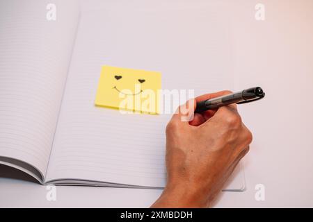 Eine Person schreibt auf ein Blatt Papier mit einem Smiley-Gesichtsaufkleber drauf. Der Aufkleber ist gelb und hat ein Herz. Die Person benutzt einen Stift, um zu wr Stockfoto