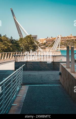 Eine moderne Brücke mit einzigartigen Designelementen, die einen ruhigen Fluss überspannt, umgeben von üppigen Bäumen und entfernten Gebäuden unter einem klaren blauen Himmel. Stockfoto
