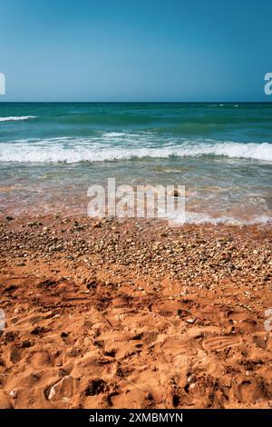 Lebhafte Strandszene mit orangefarbenem Sand, Kieselsteinen und blauen Meereswellen unter klarem Himmel. Perfekt für Sommer- und Urlaubskonzepte. Stockfoto