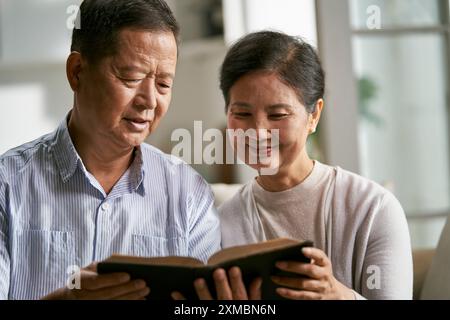 Glückliches älteres asiatisches Paar, das zu Hause auf der Familiencouch sitzt und zusammen Bibel liest Stockfoto