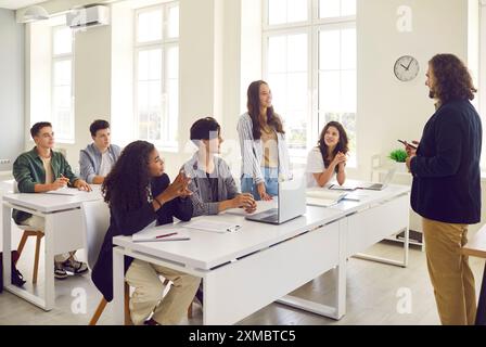 Junge Schülerin, die am Schreibtisch im Klassenzimmer steht und dem männlichen Lehrer antwortet. Stockfoto