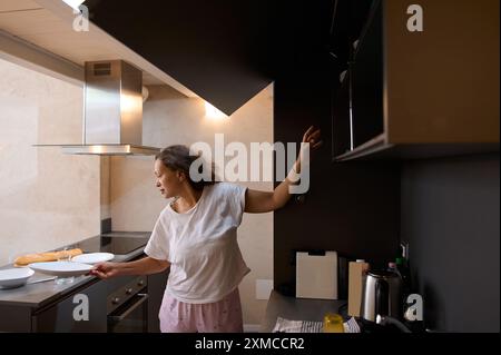 Frau in legeren Kleidern, die das Frühstück in einer modernen Küche zubereitet. Sie arrangiert Teller und Brot auf der Arbeitsplatte und fügt ein gemütliches und heimeliges Atmo hinzu Stockfoto
