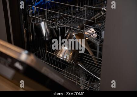 Ein offener Geschirrspüler mit verschiedenen Küchenartikeln, einschließlich Metalltöpfen, Utensilien und einem Holzlöffel. Der Geschirrspüler ist in einer modernen Küchenzeile Stockfoto