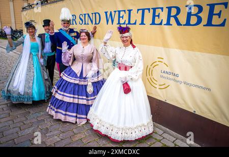 Schwerin, Deutschland. Juli 2024. Mitglieder des Schlossverbandes in historischen Kostümen begrüßen vor einem Banner mit der Aufschrift „Wir sind Weltkulturerbe“ nach der Entscheidung, das Schweriner Residenzensemble in die UNESCO-Liste des Weltkulturerbes aufzunehmen. In Neu-Delhi wurde entschieden, das Schweriner Residence Ensemble in die begehrte UNESCO-Liste des Weltkulturerbes aufzunehmen. Schwerin hofft, dass seine Aufnahme in die Liste des UNESCO-Weltkulturerbes seine touristische Attraktivität steigern und mehr internationale Aufmerksamkeit erregen wird. Quelle: Jens Büttner/dpa/Alamy Live News Stockfoto