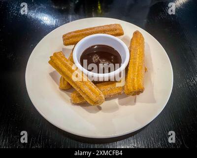 Ein Teller goldbrauner Churros mit Zucker wird mit einem kleinen Ramekin aus reichhaltiger, dunkler Schokoladensauce serviert. Die Churros sind knusprig und perfekt Stockfoto