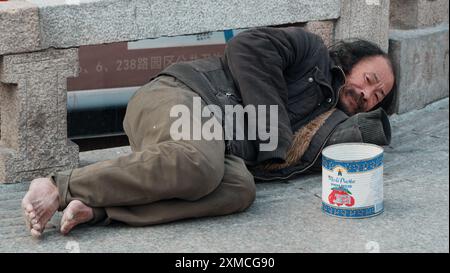 Suzhou, China - 01. Januar 2019: Ein Obdachloser liegt auf dem Bürgersteig und scheint sich an einer Steinmauer zu ruhen. Stockfoto