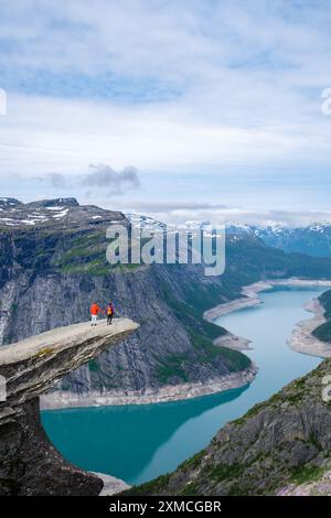 Zwei Wanderer stehen am Rande der Trolltunga Klippe in Norwegen mit Blick auf einen atemberaubenden türkisfarbenen See und eine riesige Berglandschaft. Stockfoto