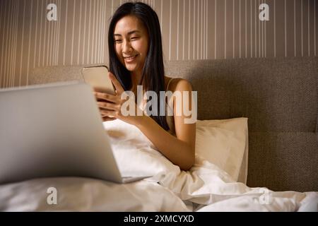 Lächelnde asiatische Frau, die auf Smartphone und Laptop im Bett sitzt Stockfoto