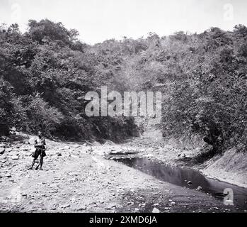 Eine Einzelfigur steht 1871 am felsigen Flussbett eines ruhigen Baches in den üppigen Wäldern von Formosa, Taiwan. Die Landschaft bietet dichte Vegetation und steile Hügel, die einen Einblick in die natürliche Schönheit der Region bieten. Das Individuum scheint die ruhige Umgebung zu beobachten, die den Entdeckungsgeist der Epoche widerspiegelt. Stockfoto