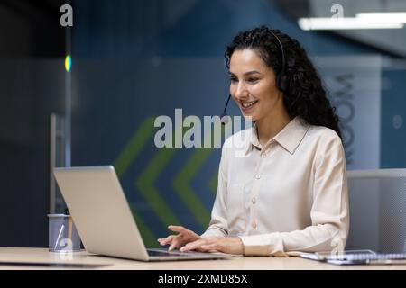 Lächelnder Kundendienstmitarbeiter, der sich mit Kunden über ein Headset in Verbindung setzt, während er ein Notebook verwendet. Moderne Büroumgebung, professionelle Kleidung, helle und einladende Atmosphäre. Stockfoto