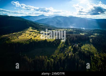 Blick aus der Vogelperspektive auf das üppige, grüne Tal mit kleinen Häusern und gewundenen Straßen, umgeben von dichten Wäldern und sanften Hügeln. Sonnenlicht filtert durch verstreute Wolken. Berge erheben sich unter blauem Himmel. Stockfoto