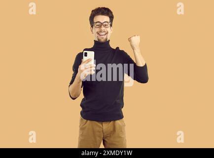 Glücklicher Mann mit schwarzem Rollkragen zeigt ja-Geste und hält Smartphone auf beigefarbenem Hintergrund. Stockfoto