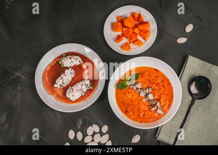 Kürbis-Karotten-Püree-Suppe mit Pfifferlingen in einer weißen Schüssel und Quark Quenelles mit Kräutern und Pfannkuchen auf einem weißen Teller auf einer Gra Stockfoto