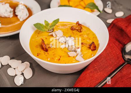 Gemüsesuppe, Kürbis- und Karottenpüree mit Samen, Pilzen, Pfifferlingen und Minze in einer weißen Schüssel auf grauem Hintergrund mit einer burgunderroten Serviette und Stockfoto