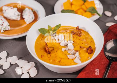 Gemüsesuppe, Kürbis- und Karottenpüree mit Samen, Pilzen, Pfifferlingen und Minze in einer weißen Schüssel auf grauem Hintergrund mit einer burgunderroten Serviette und Stockfoto