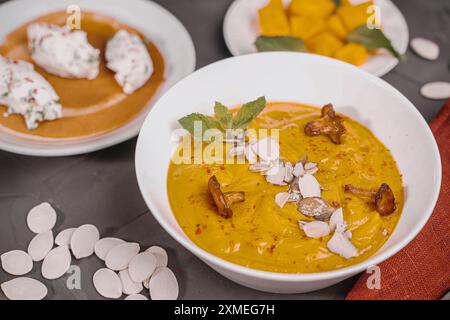 Gemüsesuppe, Kürbis- und Karottenpüree mit Samen, Pilzen, Pfifferlingen und Minze in einer weißen Schüssel auf grauem Hintergrund mit einer burgunderroten Serviette und Stockfoto