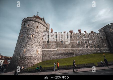 Windor Castle, Großbritannien - 16. Oktober 2023 : eine imposante Burgmauer aus Stein mit einem Turm auf der linken Seite, einem grasbewachsenen Hügel und Menschen, die auf einem Bürgersteig spazieren. Stockfoto