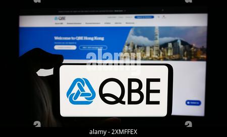 Person, die ein Mobiltelefon mit dem Logo der australischen QBE Insurance Group Limited vor der Website des Unternehmens hält. Schwerpunkt auf dem Display des Telefons. Stockfoto