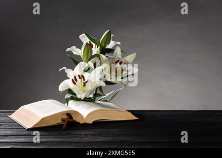 Bibel, Kreuz und Lilien auf dunklem Holztisch vor grauem Hintergrund, Platz für Text. Religion des Christentums Stockfoto