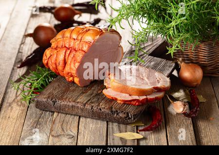 Leckerer geräucherter Schweineschinken mit Rosmarin, Zwiebeln, Knoblauch, rotem Pfeffer und Lorbeerblättern. Geräuchertes Fleisch auf einem Holztisch. Stockfoto