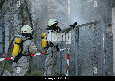 Retter in Schutzgummibezügen, die Pflanzengebiet mit Spritze bewässern. Rettungsmannstraining zur Dekontamination. Stockfoto