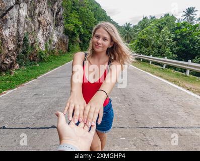 Eine junge Frau mit langen blonden Haaren, die ein rotes Oberteil und blaue Shorts trägt, hält die Hände mit dem Fotografen, während sie auf einer tropischen Straße spaziert. Stockfoto