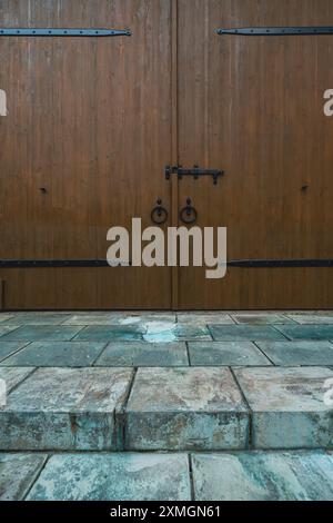 Nahaufnahme einer großen Holztür mit kunstvollen Metallgriffen und Scharnieren, die zu einem Gebäude mit alten Steinstufen im Vordergrund führt. Stockfoto