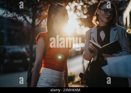 Zwei Geschäftsfrauen, die sich draußen bei Sonnenuntergang unterhalten, eine mit einem Notizbuch Stockfoto