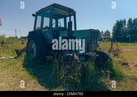 Ein alter, verrosteter Traktor, der in einem ländlichen Hinterhof mit bewachsenem Gras neben Häusern und Versorgungsmasten unter klarem blauem Himmel geparkt ist. Stockfoto