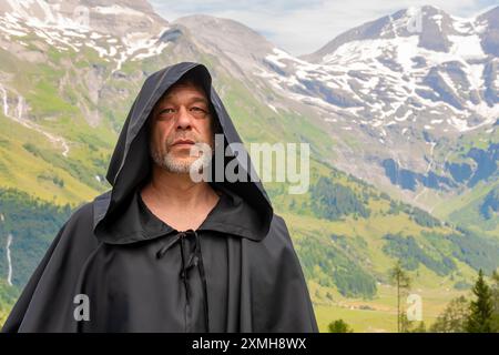 Ein bärtiger Mönch in einem Kapuzengewand steht bei Tageslicht auf einem üppigen grünen Feld an den Bergen Stockfoto