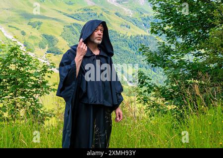 Ein bärtiger Mönch in einem Kapuzengewand steht bei Tageslicht auf einem üppigen grünen Feld an den Bergen Stockfoto