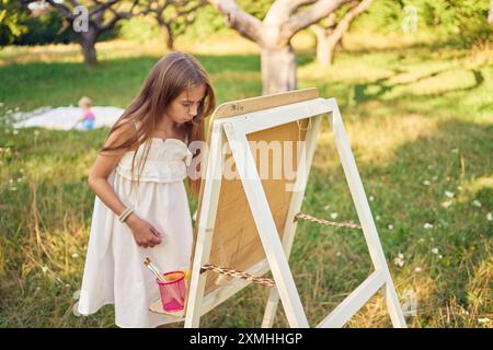 Mutter und kleine Schwester sehen zu, wie ein junges Mädchen in einem Sommerkleid ein Bild im Garten auf einer Staffelei malt Stockfoto