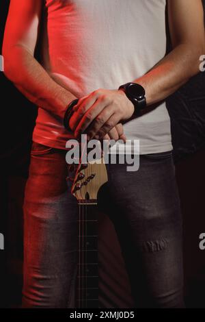 Ein Gitarrist, der mit rotem Licht beleuchtet ist und den Hals einer elektrischen Gitarre hält, die geerdet ist. Er trägt ein weißes Hemd und eine Uhr Stockfoto