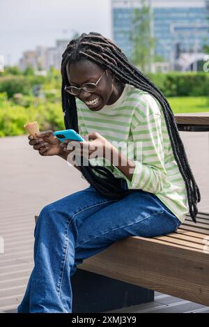 Lächelnde Afroamerikanerin mit Brille genießt Eis, während sie ihr Smartphone benutzt und auf einer Bank in einem sonnigen Park sitzt. Stockfoto