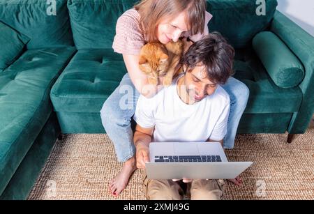 Glückliches Paar, das auf einem Sofa mit Laptop sitzt und lächelt, junges Paar, das einen Videoanruf macht, glückliche junge Familie mit einer Katze, die eine Videokonferenz veranstaltet Stockfoto