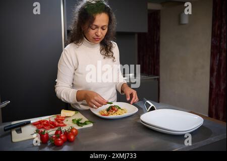 Frau in einer modernen Küche, die eine Pasta mit Basilikum garniert. Frische Zutaten wie Tomaten und Käse stehen auf der Arbeitsplatte. Stockfoto